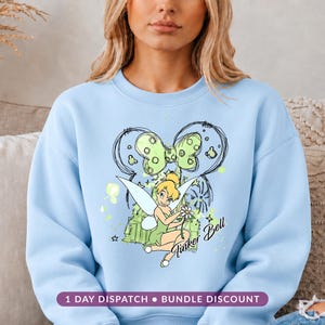 May include: A light blue crewneck sweatshirt featuring a graphic print of Tinker Bell sitting and holding a flower. Behind her is a sketch-style outline of a bow and a castle silhouette in shades of green and black. The text Tinker Bell is printed.