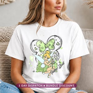 May include: A white short-sleeved t-shirt featuring a graphic of Tinker Bell sitting and holding a white flower. The design includes a large green polka-dot bow, a sketch-style castle outline, and the text Tinker Bell in a cursive font.