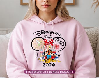 May include: A light pink hooded sweatshirt featuring a graphic of Mickey Mouse ears containing a Disneyland Paris castle, Tinker Bell, and various Disney characters. The text reads Disneyland Paris, Olivia, and 2026, with a red polka dot bow.