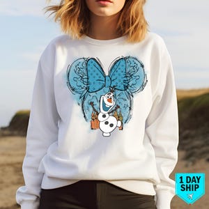 May include: White sweatshirt featuring a blue and white cartoon snowman wearing a blue bow. The snowman is in front of a blue and white castle with fireworks. The design is in a sketch style.