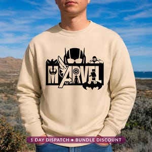 May include: A beige crewneck sweatshirt featuring a black graphic print on the chest. The design incorporates the word MARVEL in large block letters, integrated with various superhero silhouettes, symbols, and icons including a helmet and a hammer.