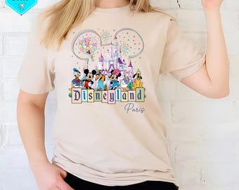 May include: A beige t-shirt with a colorful graphic of Mickey Mouse and friends in front of the Disneyland Paris castle. The graphic includes balloons, fireworks, and the text "Disneyland Paris".