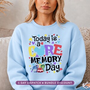 May include: A light blue crewneck sweatshirt featuring a colourful graphic print that reads Today is a CORE MEMORY Day. The design includes a blue silhouette of mouse ears, small star accents, and five small character figures at the bottom.