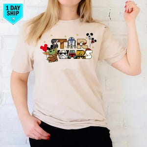 May include: A beige t-shirt featuring a graphic of Star Wars characters, including Yoda, R2-D2, Darth Vader, and Mickey Mouse. The text "STAR WARS" is written in a colourful font.