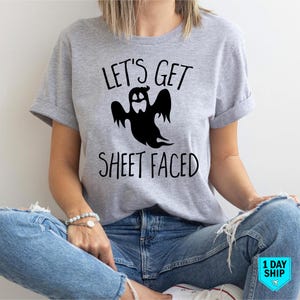 May include: A light grey t-shirt featuring the text "LET'S GET SHEET FACED" and a black ghost illustration. The shirt has short sleeves and is worn with blue jeans. The design is suitable for Halloween.