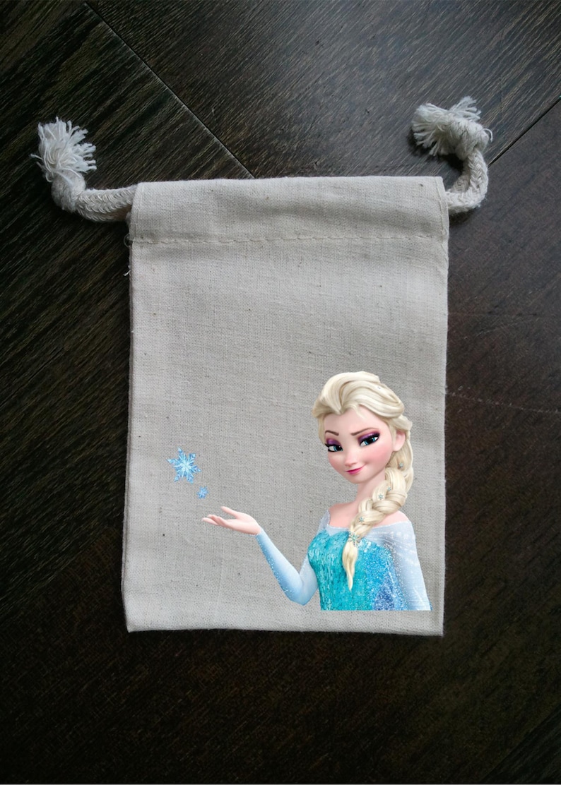 Puede incluir: Una bolsa de tela blanca con cord&oacute;n de ajuste, impresa con una imagen de una princesa Disney de cabello rubio con un vestido azul y extendiendo la mano. La princesa est&aacute; rodeada de copos de nieve.