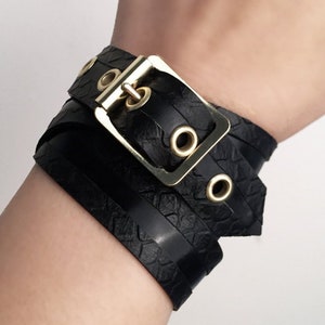 May include: A black leather bracelet with a gold buckle and multiple straps. The bracelet has a textured, snake-like pattern.