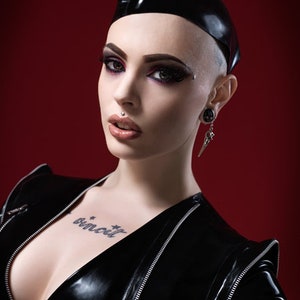 May include: A woman wearing a black leather jacket with a zipper detail and a black leather cap. She has a tattoo on her chest that says "binecuit".