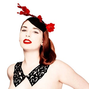 May include: A woman wearing a black lace collar and a black hat with red flowers. The hat is tilted to the side and the flowers are arranged in a cluster.