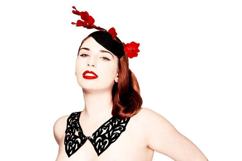 May include: A woman wearing a black lace collar and a black hat with red flowers. The hat is tilted to the side and the flowers are arranged in a cluster.