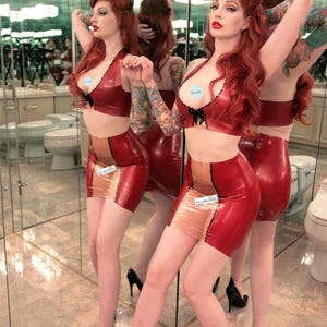 May include: A woman with long red hair is wearing a red latex bra and skirt with a nude panel. She is standing in a bathroom with mirrors and has tattoos on her arms.