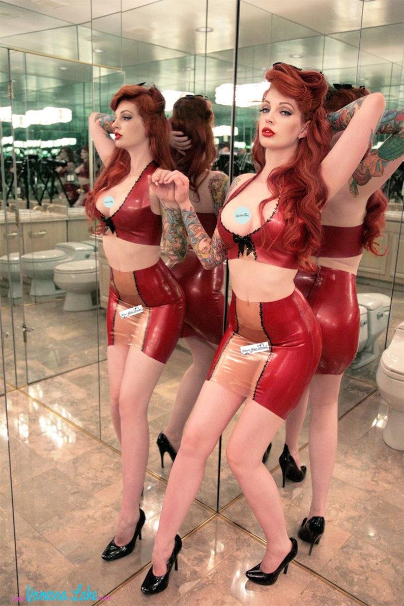 May include: A woman with long red hair is wearing a red latex bra and skirt with a nude panel. She is standing in a bathroom with mirrors and has tattoos on her arms.