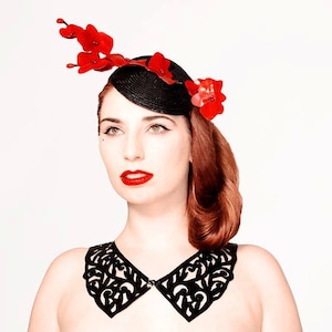 May include: A woman wearing a black leather collar with a delicate cut-out design and a black hat with red flower accents.