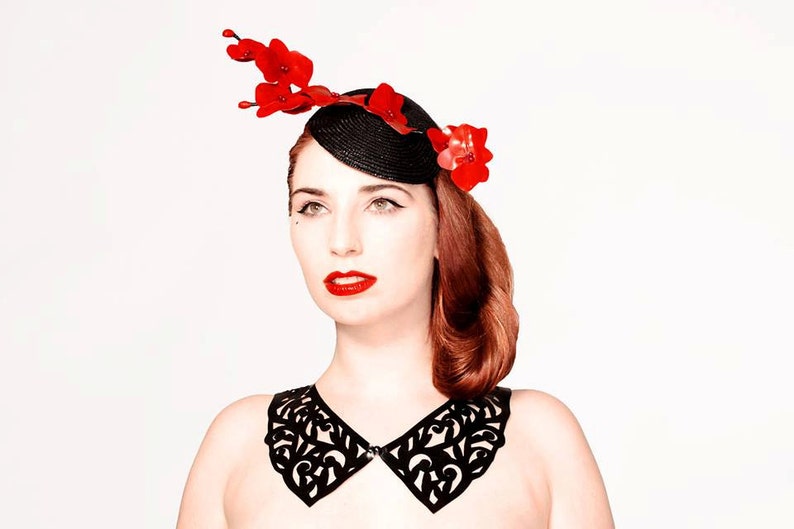 May include: A woman wearing a black leather collar with a delicate cut-out design and a black hat with red flower accents.