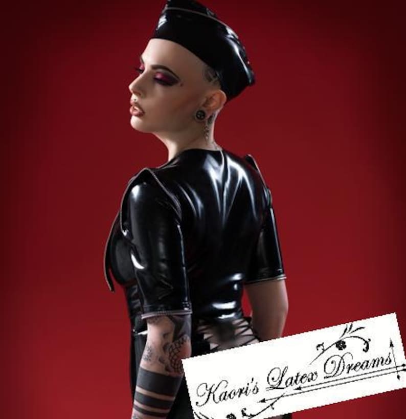 May include: A woman wearing a black latex top and a black cap. The top has a short sleeve and a zipper on the back. The woman has tattoos on her arm. The image has the text "Kaori's Latex Dreams" in the bottom right corner.