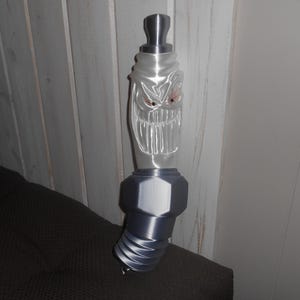 May include: A gray and clear decorative spark plug with a menacing face design. The spark plug has a silver top and base, with a clear, frosted glass-like center featuring a carved face with red eyes and a wide grin.