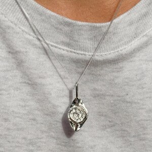 Könnte beinhalten: Halskette mit einem silbernen Anhänger in Tropfenform. Der Anhänger hat eine strukturierte Oberfläche und ein Spiralmuster in der Mitte. Er hängt an einer feinen Silberkette. Präsentiert vor grauem Hintergrund, schlicht und elegant.