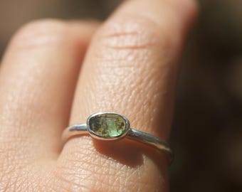 May include: A silver ring with an oval, olive-green gemstone. The ring has a thin, polished band, and the gemstone is set in a bezel. The ring is displayed on a finger, with a blurred background.