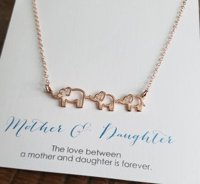 Puede incluir: Un collar de oro rosa con tres siluetas de elefantes conectadas. El collar est&aacute; sobre una tarjeta blanca con el texto "Madre e Hija. El amor entre una madre y su hija es para siempre."