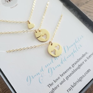 Puede incluir: Tres collares de oro con colgantes en forma de corazón. El colgante más grande tiene dos corazones recortados. Los collares se muestran en una tarjeta blanca con el texto "Grandmother & Granddaughter" y un mensaje sobre el amor entre abuela, hija y nieta.