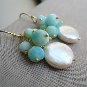 May include: A pair of drop earrings featuring clusters of round, light blue stones and a single, flat, circular white pearl attached to gold-toned metal hooks. The stones vary slightly in size, creating a textured, layered appearance on each earring.