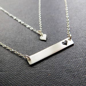 May include: A silver-colored necklace with two strands. The longer strand features a rectangular bar pendant with a heart-shaped cutout. The shorter strand has a small heart pendant. The jewelry is displayed on a dark textured surface.