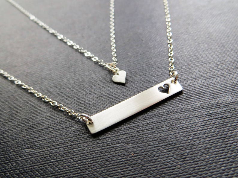 May include: A silver-colored necklace with two strands. The longer strand features a rectangular bar pendant with a heart-shaped cutout. The shorter strand has a small heart pendant. The jewelry is displayed on a dark textured surface.