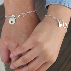 Könnte beinhalten: Zwei silberne Unendlichkeitsarmbänder mit Herzanhängern. Ein Armband hat einen Herzanhänger mit einem ausgeschnittenen Herzen, das andere einen massiven Herzanhänger.