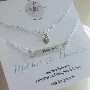 May include: A silver necklace set featuring two delicate chains. One chain has a small heart pendant, and the other has a bar pendant engraved with the name "Evelyn" and a heart cutout. The card reads "Mother & Daughter".