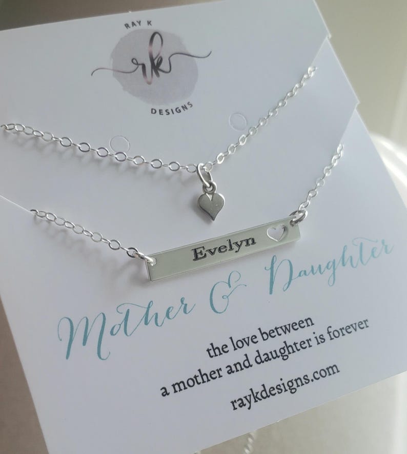 May include: A silver necklace set featuring two delicate chains. One chain has a small heart pendant, and the other has a bar pendant engraved with the name "Evelyn" and a heart cutout. The card reads "Mother & Daughter".