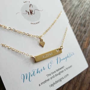 May include: Two gold necklaces on a card that reads "Mother & Daughter." The top necklace has a small heart pendant. The bottom necklace has a bar pendant with the name "ALEENA" engraved and a heart cutout. The card also has the text "The love between a mother and daughter is forever."