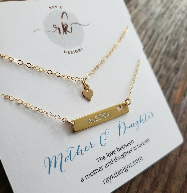May include: Two gold necklaces on a card that reads "Mother & Daughter." The top necklace has a small heart pendant. The bottom necklace has a bar pendant with the name "ALEENA" engraved and a heart cutout. The card also has the text "The love between a mother and daughter is forever."
