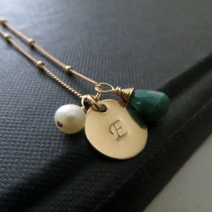 May include: Gold necklace featuring a small pearl, a green gemstone, and a gold charm engraved with the letter "E".