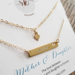 May include: A gold-colored necklace set with two chains. One chain has a small heart pendant, and the other has a rectangular bar engraved with the name "ALEENA" and a heart cutout. The card reads "Mother & Daughter".