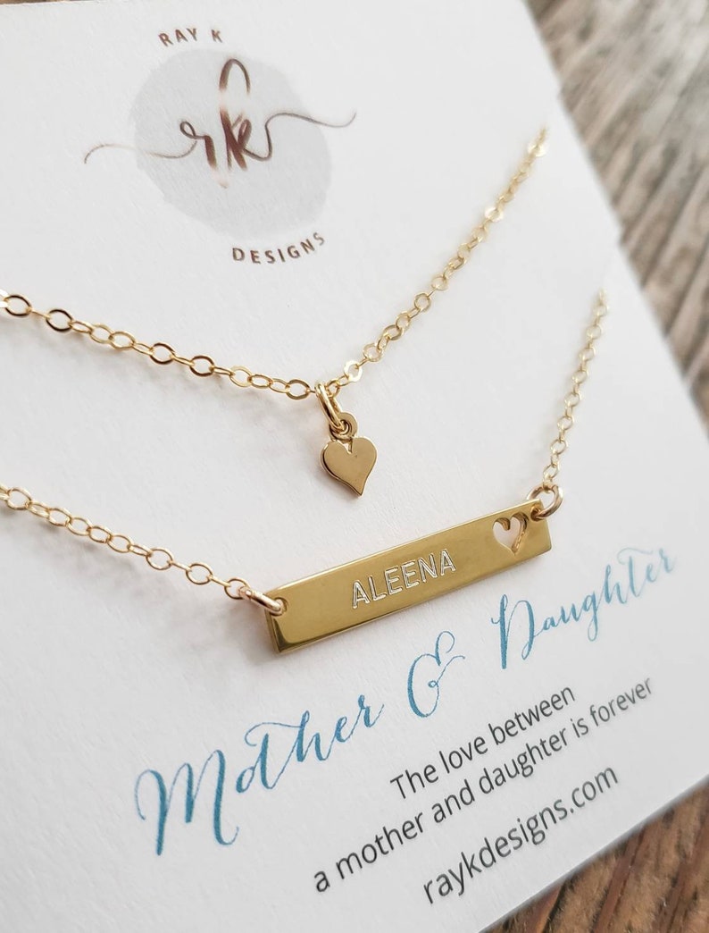 May include: A gold-colored necklace set with two chains. One chain has a small heart pendant, and the other has a rectangular bar engraved with the name "ALEENA" and a heart cutout. The card reads "Mother & Daughter".