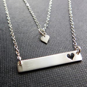 May include: A silver-toned necklace set featuring two chains. The longer chain has a rectangular bar pendant with a heart cutout. The shorter chain has a small heart pendant. The jewelry is displayed on a dark textured surface.