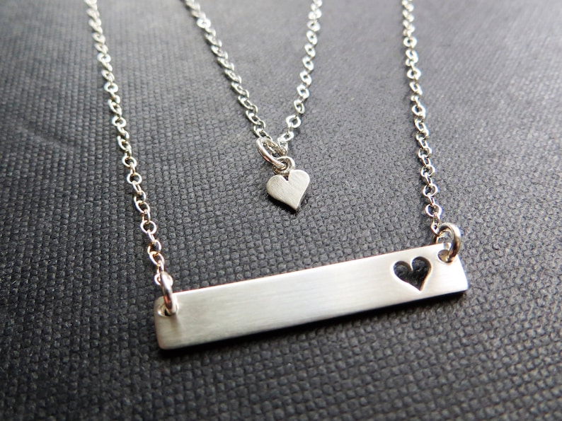 May include: A silver-toned necklace set featuring two chains. The longer chain has a rectangular bar pendant with a heart cutout. The shorter chain has a small heart pendant. The jewelry is displayed on a dark textured surface.