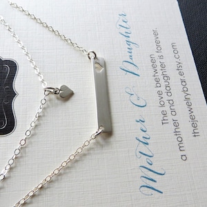 May include: A silver necklace with a rectangular pendant and a small heart charm. The necklace is on a card with the text "Mother & Daughter" and "thejewelrybar.etsy.com". The necklace is a delicate piece of jewelry.