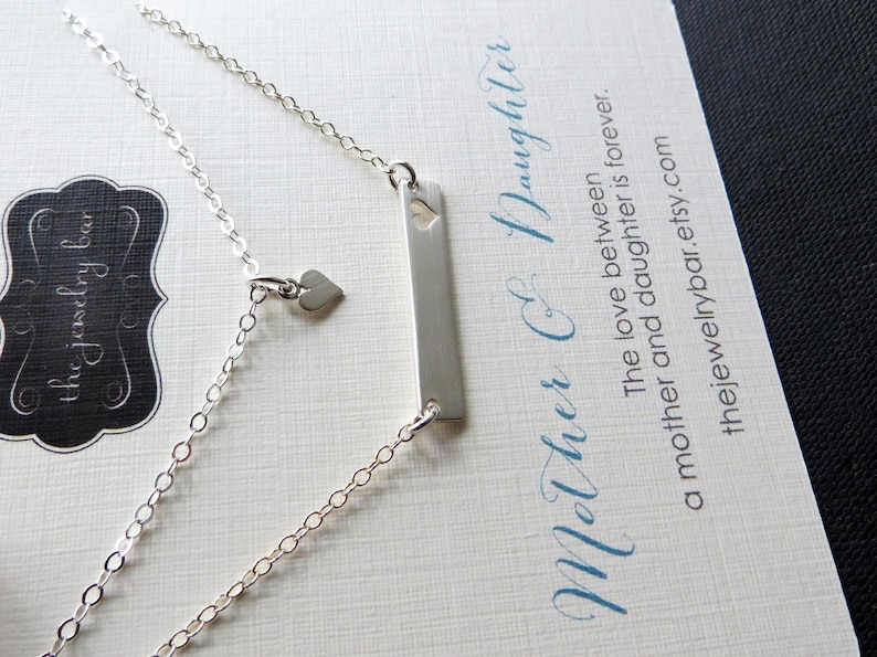 May include: A silver necklace with a rectangular pendant and a small heart charm. The necklace is on a card with the text "Mother & Daughter" and "thejewelrybar.etsy.com". The necklace is a delicate piece of jewelry.
