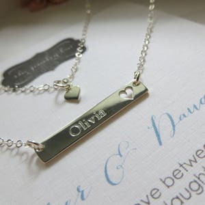 May include: A silver-colored necklace with a rectangular pendant engraved with the name "Olivia" and a heart-shaped cutout. The necklace has a delicate chain and a small heart charm near the clasp. The background includes a card with cursive text.