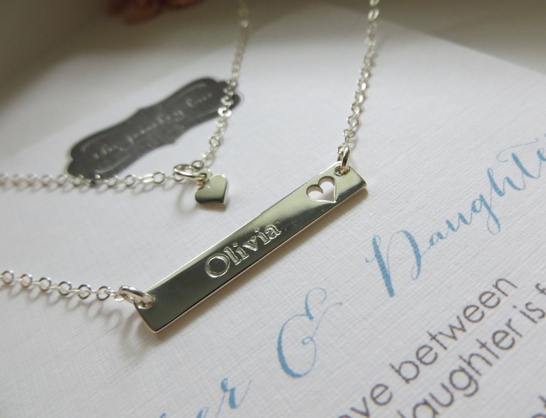 May include: A silver-colored necklace with a rectangular pendant engraved with the name "Olivia" and a heart-shaped cutout. The necklace has a delicate chain and a small heart charm near the clasp. The background includes a card with cursive text.