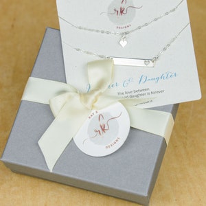 May include: A silver necklace set with a heart pendant and a bar pendant with a heart cutout, displayed on a card that reads "Mother & Daughter." The set is presented in a gray gift box tied with a cream ribbon and a circular tag with the brand logo.