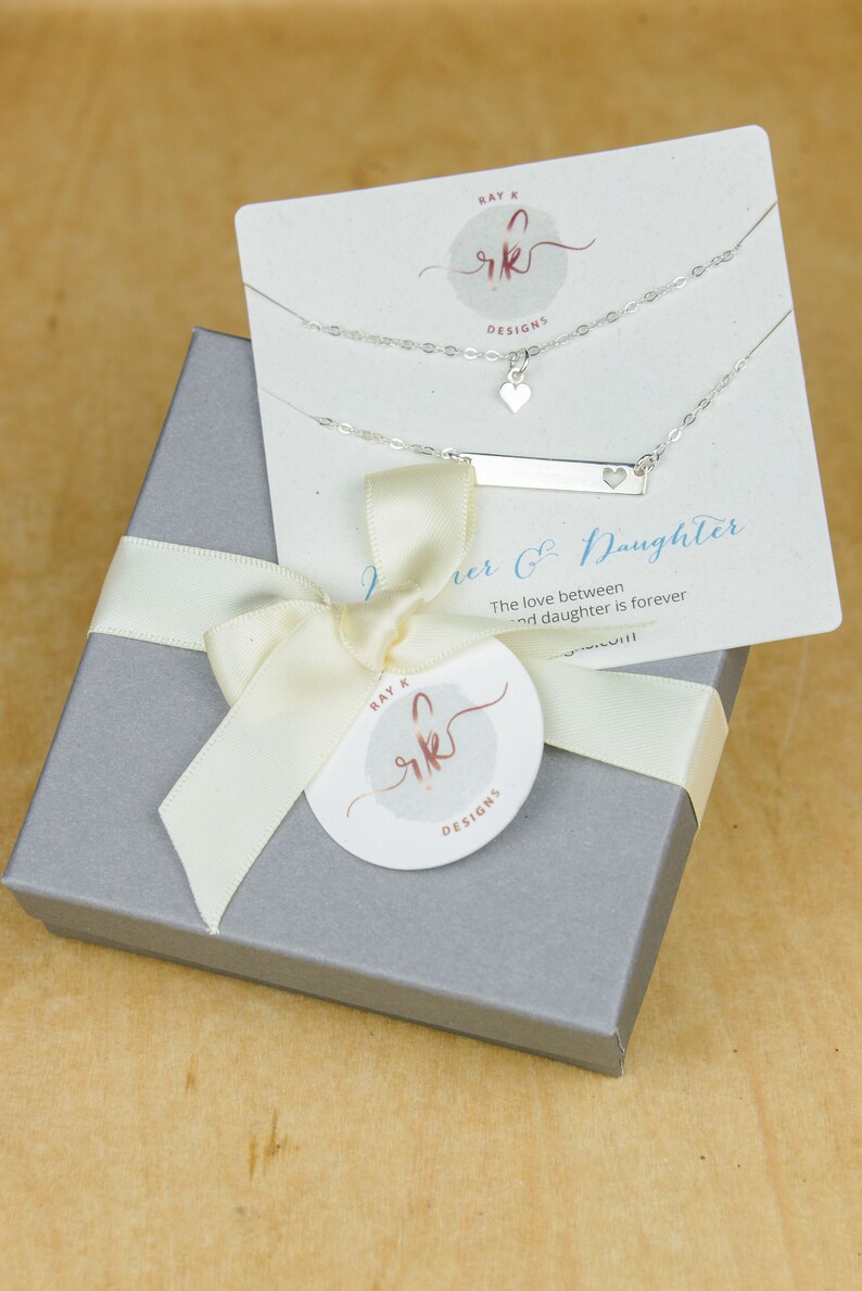 May include: A silver necklace set with a heart pendant and a bar pendant with a heart cutout, displayed on a card that reads "Mother & Daughter." The set is presented in a gray gift box tied with a cream ribbon and a circular tag with the brand logo.