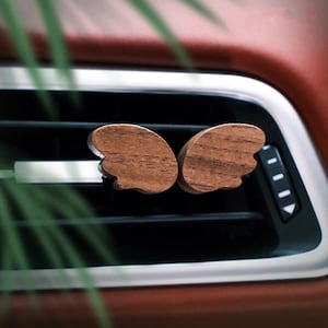 May include: Two wooden wing-shaped car vent clips. The clips are made of a dark brown wood and are attached to the car's vent. The background shows a red interior and a silver vent.