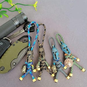 Op de afbeelding: Vier handgemaakte Spartaanse krijger sleutelhangers in blauw, groen en bruin paracord. Elke sleutelhanger heeft een bronzen helm en schild. Een zaklamp, sleutels en een zakmes staan op de achtergrond.