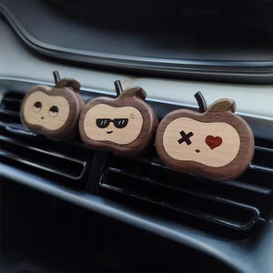 Puede incluir: Tres ambientadores para coche con forma de manzana de madera con diferentes expresiones faciales. Las manzanas son marrones con un centro de madera más claro. Uno tiene gafas de sol, otro un corazón y el último una expresión de sorpresa. Se adjuntan a una rejilla de ventilación.