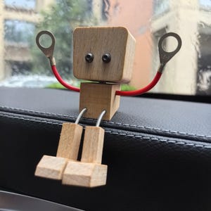 May include: A handcrafted wooden robot figurine with a square head and body, featuring black eye screws and red wire arms. The legs are made of small wooden blocks connected by silver wire. The robot is sitting on a black surface.