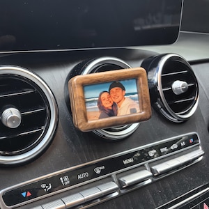 Könnte beinhalten: Ein kleiner, rechteckiger Holzfotorahmen mit einem Farbfoto von zwei Personen. Der Rahmen ist an einer Lüftungsöffnung eines Autos befestigt. Der Innenraum des Autos weist schwarze und silberne Akzente auf.