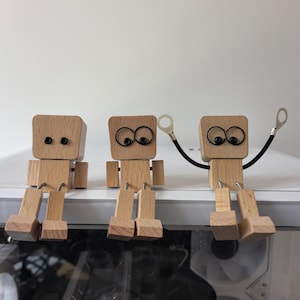 May include: Three wooden robot figures with movable limbs and large, round eyes. The robots are made of light-colored wood and are sitting on a white surface. One robot has a black wire with a silver ring attached to its arm.