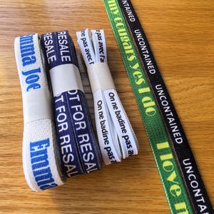 Puede incluir: Varios rollos de cinta de tela con varios textos impresos. Las cintas son blancas, azul marino y verdes, con texto en azul, blanco y amarillo. El texto incluye frases como "Emma Joe", "Resale" e "I love n".